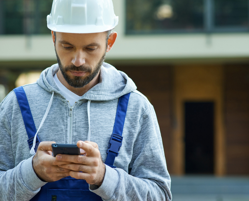 Young male builder in hardhat using smartphone