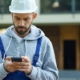 Young Male Builder In Hardhat Using Smartphone