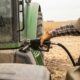 A Farmer Wearing A Plaid Shirt And Cap Refuels A Large Green Tractor With A Fuel Nozzle In A Dry Field